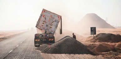 A truck unloading gravel on a dusty desert road, with a worker standing nearby and piles of construction material around